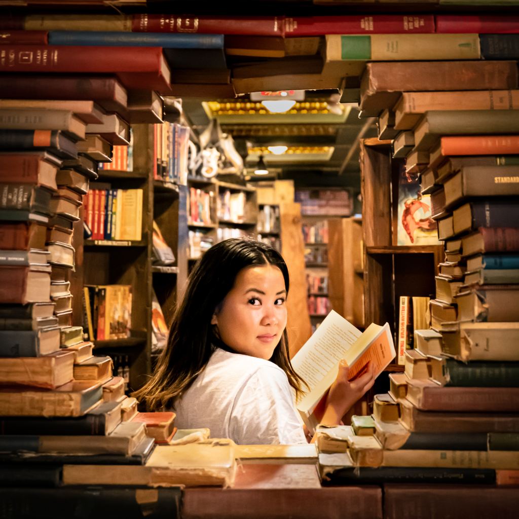 A woman in a library with long black hair holding a book and her head turned towards the viewer. She is looking through a circular gap in the books. 