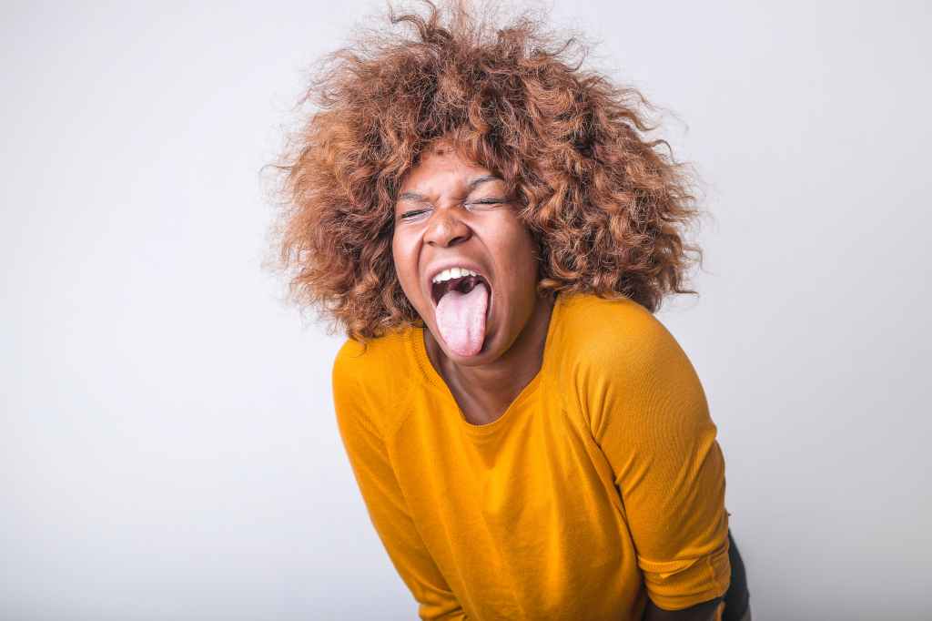 woman with yellow jumper and brown curly hair is leaning forward scrunching up her eyes and sticking her tongue out