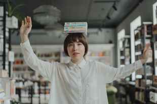 ATAR. woman balancing books on her head with arms spread. She is of asian appearance and wearing a white shirt.