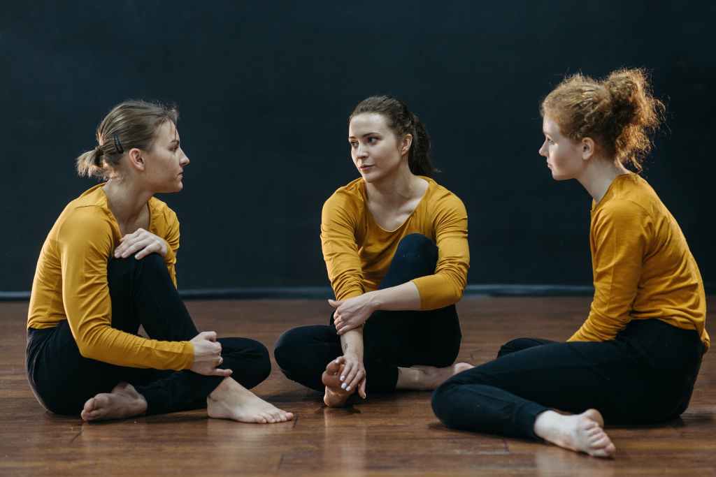Three female dancers are sitting and talking on stage about their career lessons, wearing gold jerseys and dark pants.