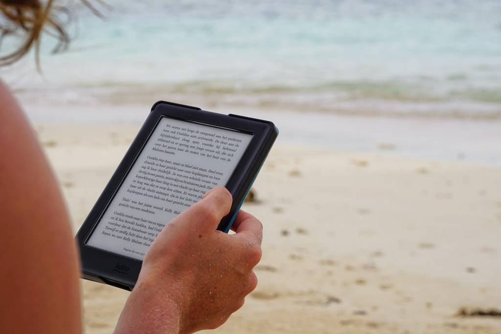 reading. Image is the shoulder arm and hand of a young woman on a beach reading a book on an e-reader