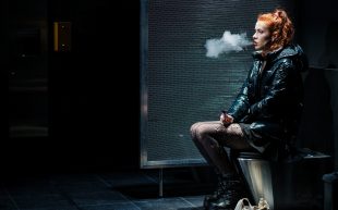 Janet Anderson as Rosie in 'Overflow'. Photo: Robert Catto. A woman with a red ponytail smoking and sitting on a toilet. She is wearing a black jacket with skirt, stockings and platform boots.