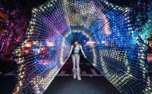 EOIs open for Un[contained] Arts Festival 2024. Photo: Supplied. A performer wearing a light device that spans from her back like a peacock tail with vibrant colourful lights. She is standing in the middle of a road at night.