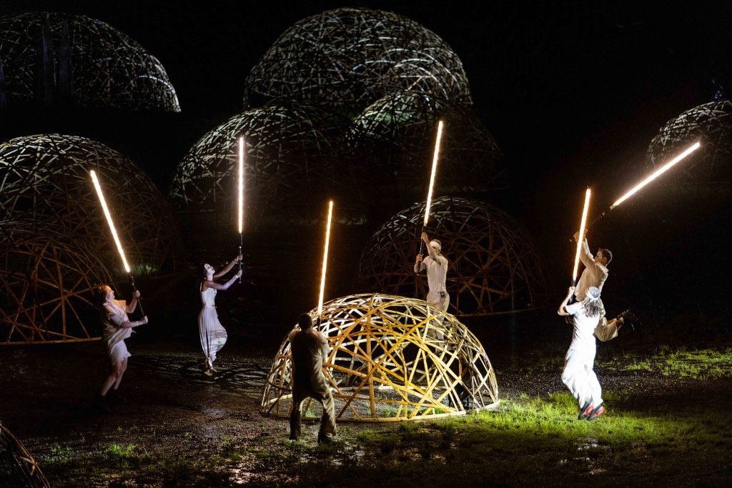 ‘The Renewal: A Village Dreams’. Photo: Courtesy of Woodford Folk Festival and Lachlan Douglas. A group of performers wearing white cluster around a woven dome, holding poles of light above them.