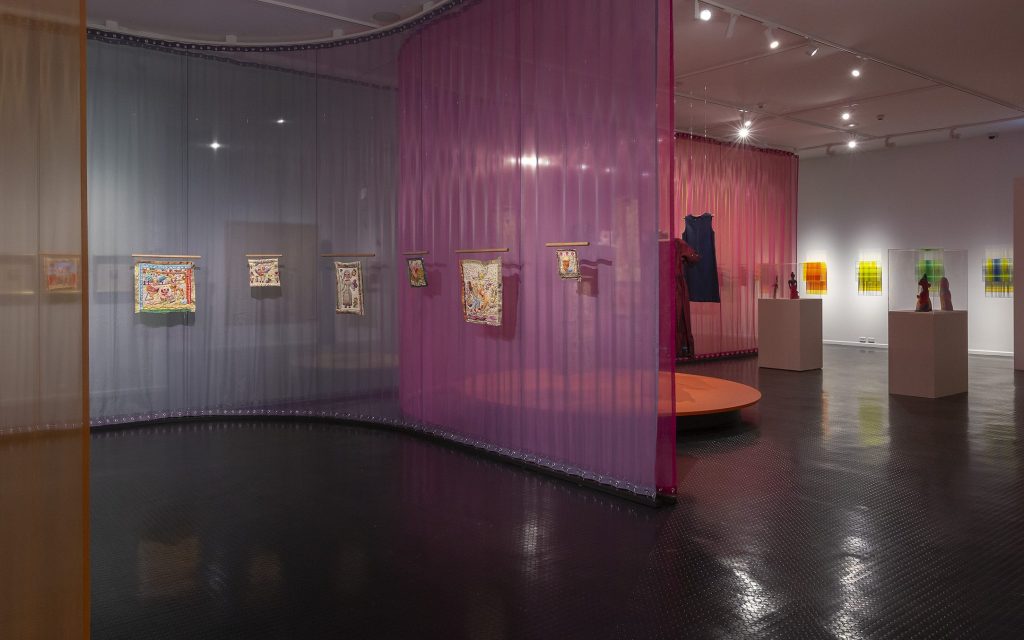 LISA GORMAN + MIRKA MORA, installation view at Warrnambool Art Gallery. Photo: Supplied. An exhibition space partitioned with orange, dark turquoise and pink sheer curtains. In front of the curtains hang embroideries by Mirka Mora and clothing by Gorman x Mirka.