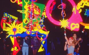 Immersive exhibition ‘130BPM: Soundtrack to a Revolution’ curated by Bertie Blackman to open at Qtopia Sydney in its new home. Image: 1985 Sydney Gay Mardi Gras, cropped with colour enhancement. Photo: William Yang. Large puppet-like pieces with vibrant colours dance in the dark with people holding them up from the bottom.