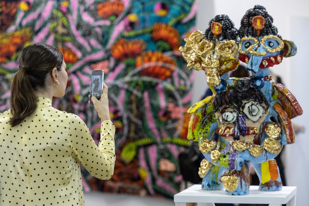 Work by Ramesh Mario Nithiyendran (Sullivan+Strumpf) at ART SG 2024. Photo: Supplied. A woman at the ART SG art fair taking a photo in front of a mid-sized ceramic sculpture. The Sculpture is colourful and looks like two adjoined elephants standing on their hind legs.