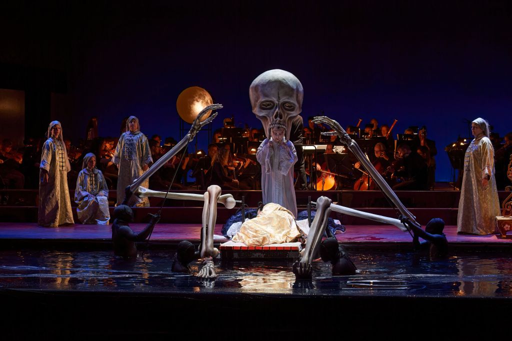 Ex Machina tells ArtsHub that 'changes have been made to Ex Machina's previous version of Stravinsky's opera, such as modifications made to the makeup'. Photo: Michael Cooper. Production photo of ‘The Nightingale’ , featuring a large skull puppet and performers standing near a body of water with the moon shining behind, as if in ritual. 