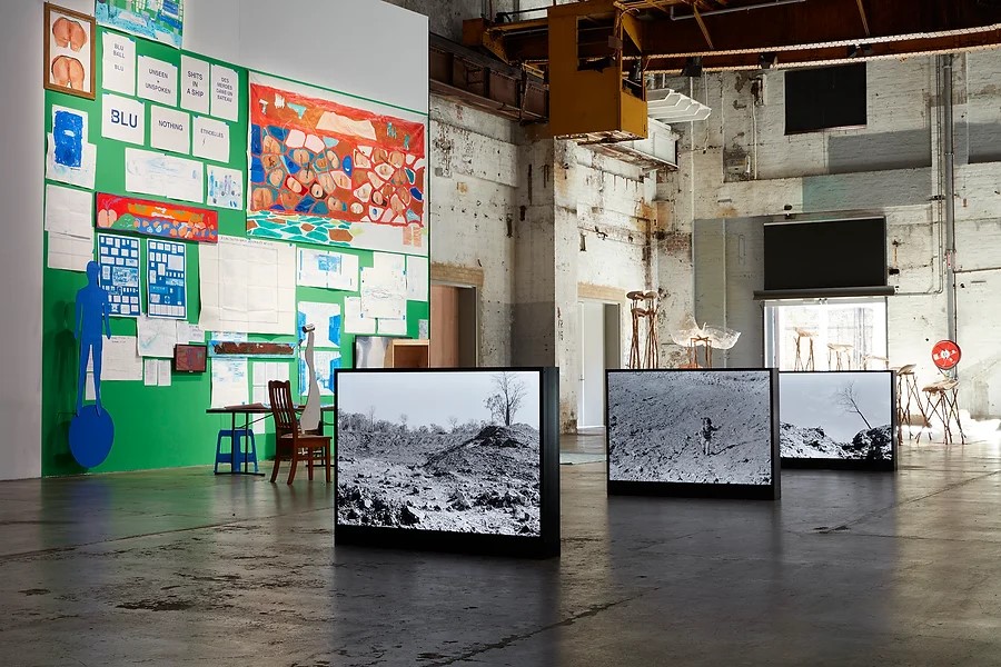 Alana Hunt, 'All the violence within / In the national interest (2019-21)', installation view at Carriageworks. Photo: Courtesy of the artist. Installations in an industrial space. One the floor are three illuminated boxed with black and white photographs of a barren landscape. On the wall is an installation with different sized posters against a green background. 