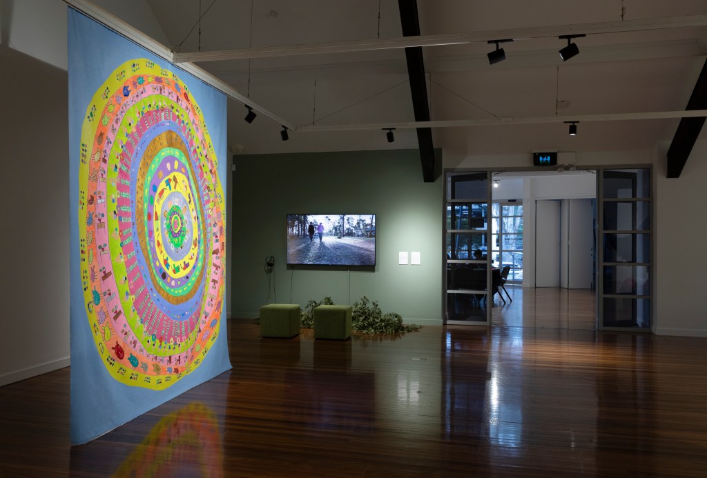 Exhibition space with dim lighting displaying a large hanging work to the left side with vibrant layers of circles and drawings. In the background is a video work showing two people standing outdoors under the sun. 