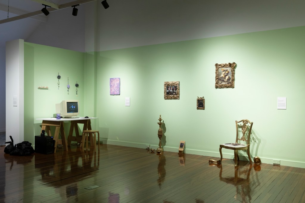 Exhibition space with light pistachio coloured walls. Furniture including a table with a box computer, an ornate chair,  a lamp post are visible. Several small paintings with elaborate gold frames hang on the wall. 