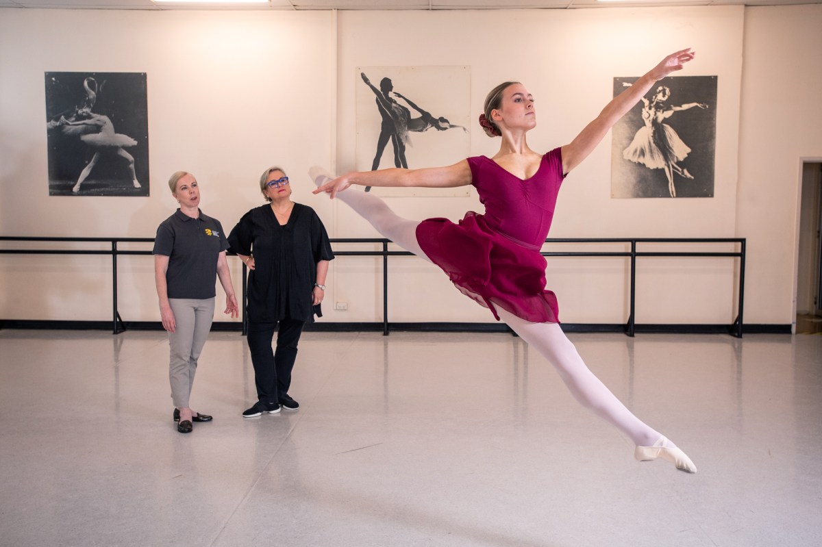 New research will source more information on dance injury. Image is two women at the back of a studio in front of the barre, watching a woman in red in mid balletic leap.