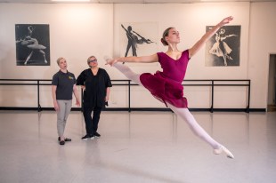 New research will source more information on dance injury. Image is two women at the back of a studio in front of the barre, watching a woman in red in mid balletic leap.