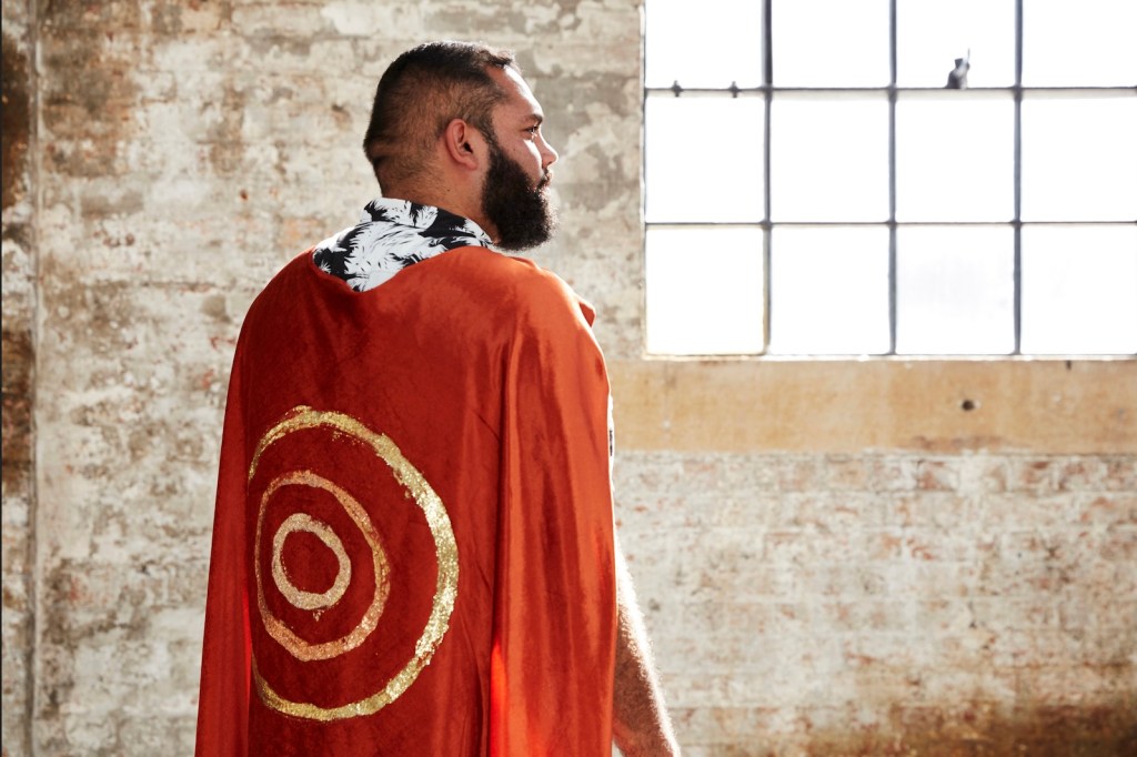 Aboriginal man with beard wearing a red cape with gold target painted. Dennis Golding.