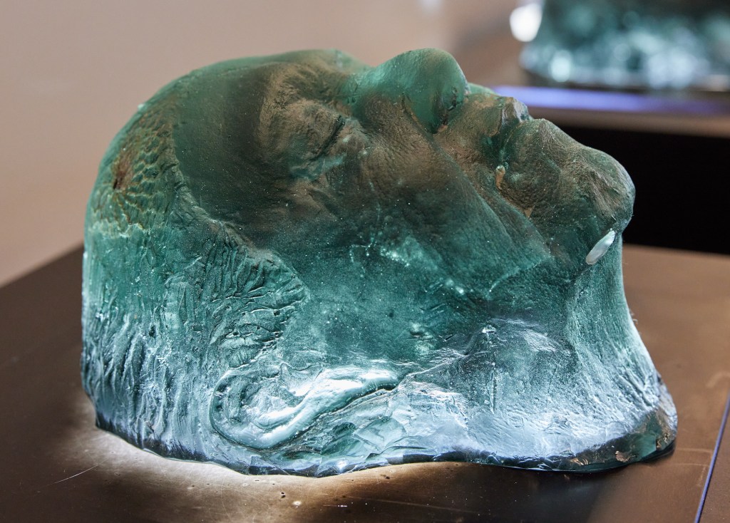 Jennifer Kemarre Martiniello OAM, 'Not Wasted', 2023. Photo: Supplied. Sculpture of a person's head in relief made from recycled television screen. The face looks peaceful with closed eyes and a side profile. The glass is illuminated from blue and is turquoise in colour.