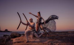 Adelaide Festival. Two First Nations performers at dusk on a beach next to the sea gesticulate, one with her arm and the other with two large boomerangs.