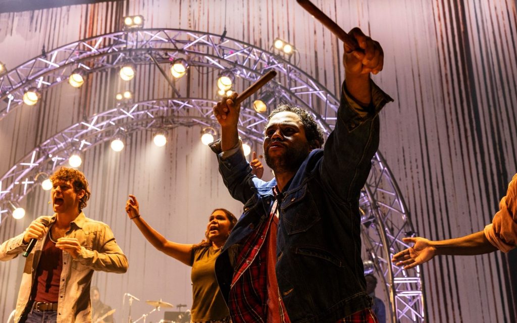 ‘Big Name, No Blankets’ premiered at Sydney Festival 2024. Photo: Brett Boardman. Close up of some of the cast of Big Name No Blankets, a man in front has his arms up holding clapsticks, bright stage lights in the background give the feeling of an epic rock show.