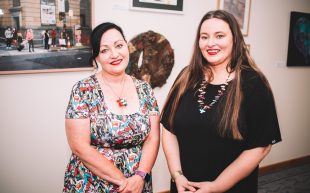 University of Southern Queensland Higher Degree Research PhD candidate Lisa Hobbs (left) with School of Creative Arts Associate Professor Beata Batorowicz (right). Photo: Supplied. Two women standing in front of a wall of artworks, smiling at the camera.