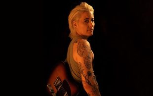 Jess McAvoy’s ‘The Search’ performing as part of Midsumma Festival 2024. Photo: Jen Macchiarelli. McAvoy (short blond hair, tattoos covering their arm and wearing a guitar behind their back) looks towards the camera, standing against a back background with soft lights.