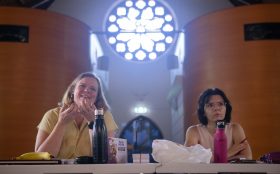 Creative development sessions for ‘Portaloo’ with L-R: Jennifer Piper and Rachel Edmonds. Photo: Sarah Clarke. In front of a high church-like round window, two women sit behind a table looking at the camera. They are watching something or someone and have snacks on the table in front of them. One on the right with short dark hair and glasses holds a pen. One on the left with medium length blonde hair is laughing.