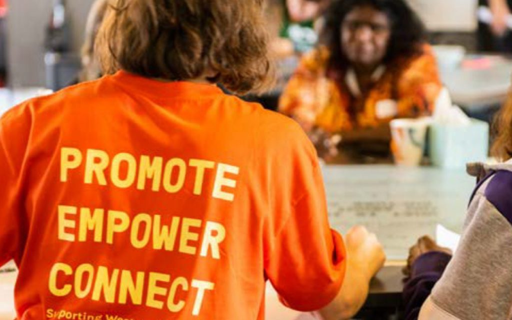 AACHWA’s Our Business: Aboriginal Art Centre Forum, Jambinu (Geraldton), 2022. Photo: Dragonfly Media. A figure with their back turned to the viewer, wearing an orange shirt with the words ‘PROMOTE EMPOWER CONNECT’.