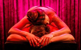 āA Body at Workā. Photo: Darren Gill. Two naked bodies lie on top of each other on a simple massage bed with their heads facing the viewer. A red light is illuminating their bodies.