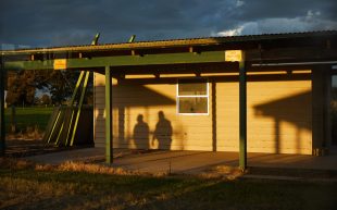 Work by Tace Stevens showing as part of group exhibition ‘Only the future revisits the past’ at Centre for Contemporary Art as part of PHOTO2024. Image: Supplied. Photography work that captures a residential shed in the glow of early evening sunset. The shadow of two figures are reflected onto the wall of the building.