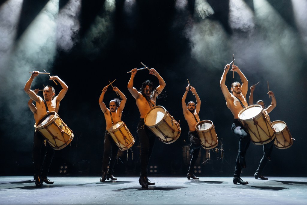 Malevo. A line-up of five bare-chested males raise their drumsticks above their heads and over the drums attached in front of them.