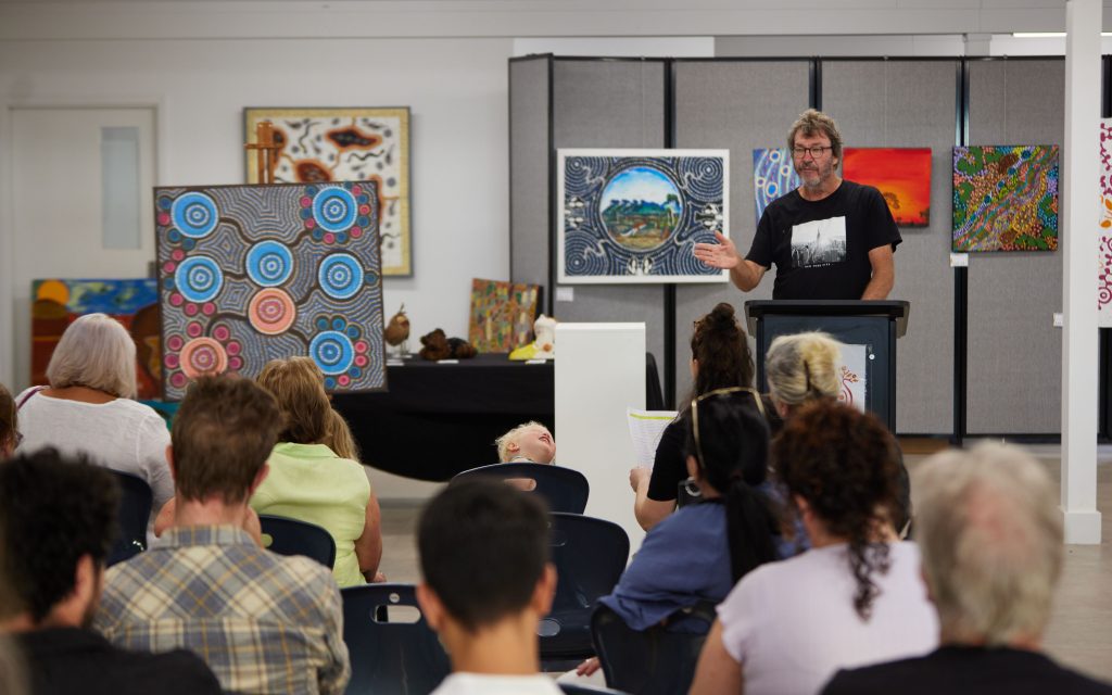 KAPP judges feedback sessions. Photo: Travis Hayto. People sitting in a room with a man standing at the podium pointing to a painting next to him. The room is filled with other artworks, mostly landscape.