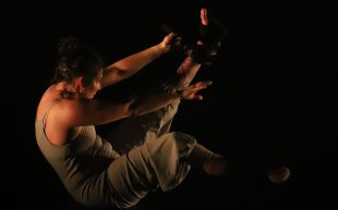 Aerialist Lauren Watson in a scene from 'NERVE'. Photo: Cinnamon Smith. A performer with her leg and arms raised, balancing on her sitting bones. The background is pitch black with soft lights illuminating her body.