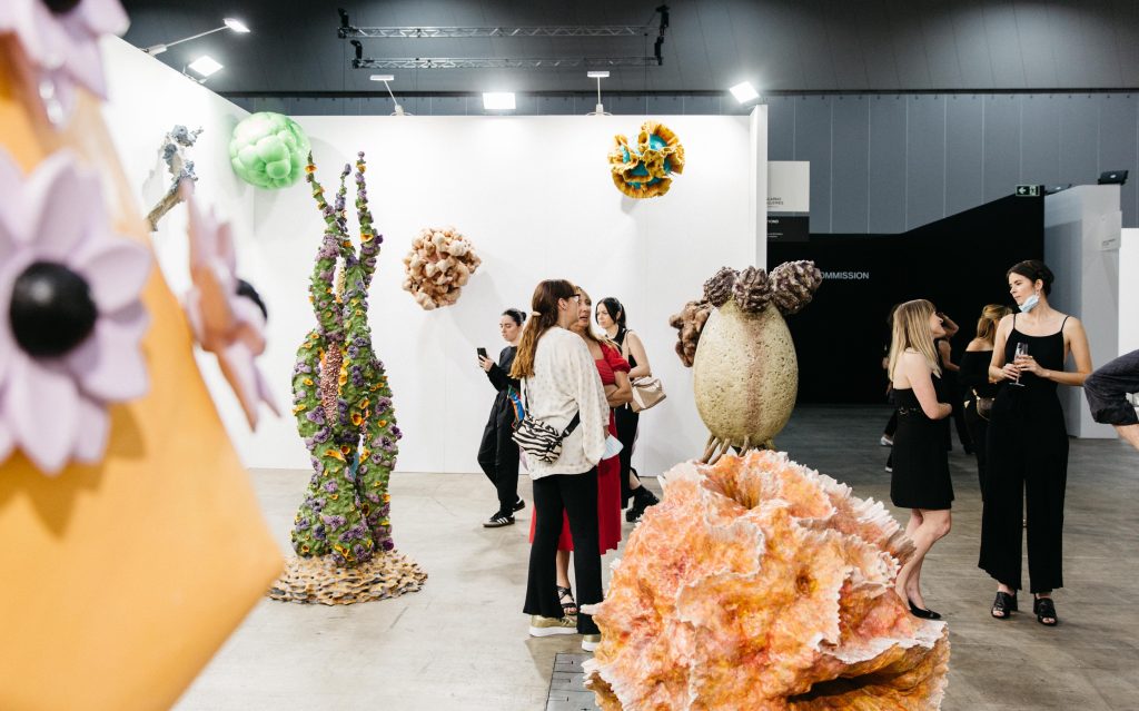 Melbourne Art Fair 2022. Photo: Marie-Luise Skibbe. People standing inside the Melbourne Convention and Exhibition Centre surrounded by artworks. Seen in the photo are installations that look like weird colourful organisms.