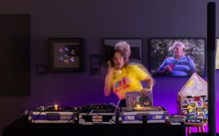 Foxy La Fuzz by Kayapa Creative Studios, presented as part of Creative Open. Now calling for artists and creatives to participate. Photo: Supplied. A figure holding a yellow phone against her ear while standing in front of a DJ set. She appears to be caught in mid-motion.