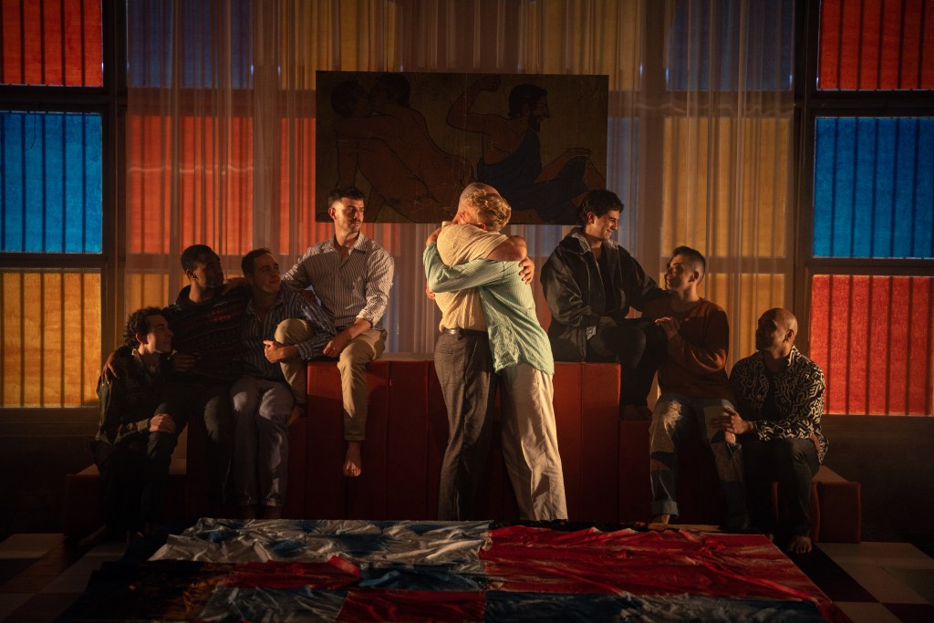 The Inheritance. A group of men on a dimly lit stage, with two spotlit in the centre hugging.