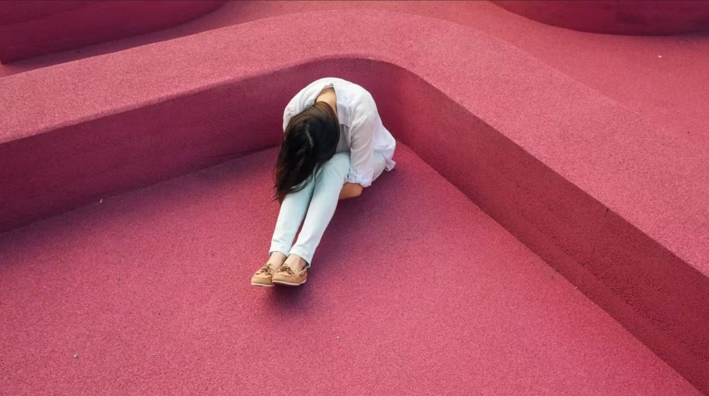 Photo: Verne Ho via Unsplash. A figure with the body bent and hugging the legs, sitting on a pink carpet.