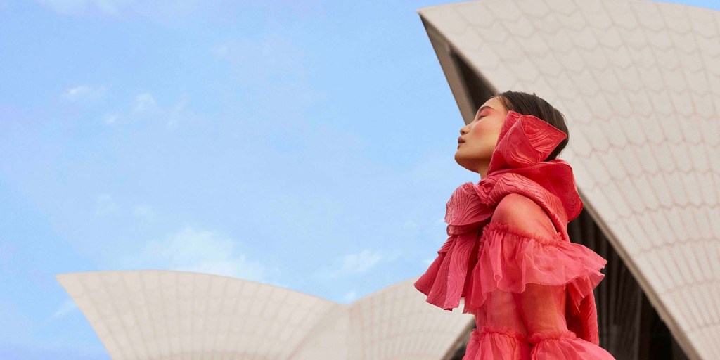 A promotional image for Opera Australia. Against the sails of the Sydney Opera House a figure in red looks up to the sky.