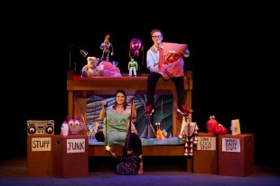 Pillow Fight. Against a black backdrop man and woman sit on a bunk bed surrounded by toys and puppets. Beside the bunk are boxes with labels like ’stuff’, ‘junk’, ‘Shane’s cool stuff’ and ‘Nadia’s baby stuff’.