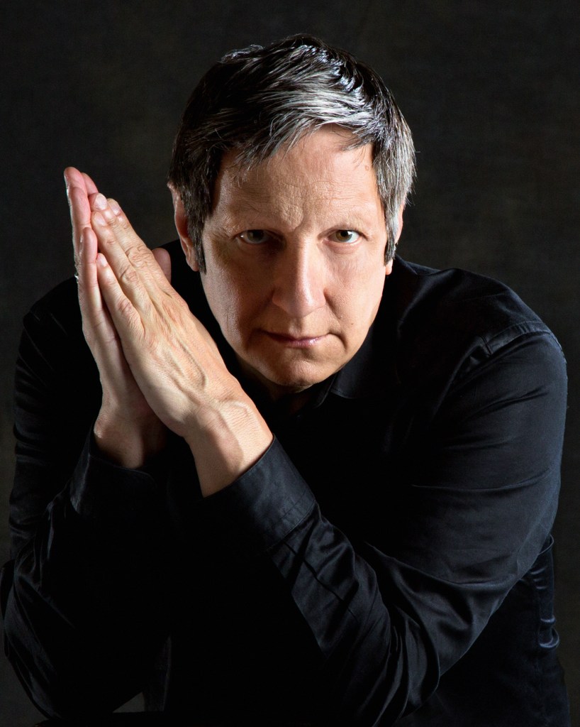 Portrait of Robert Lepage, a Canadian playwright and President of Ex Machina. Photo: Tony Hauser. He is a white man with short black and grey hair, looking intensely at the viewer. His hands are placed together and raised next to his face. He is wearing a black shirt. 