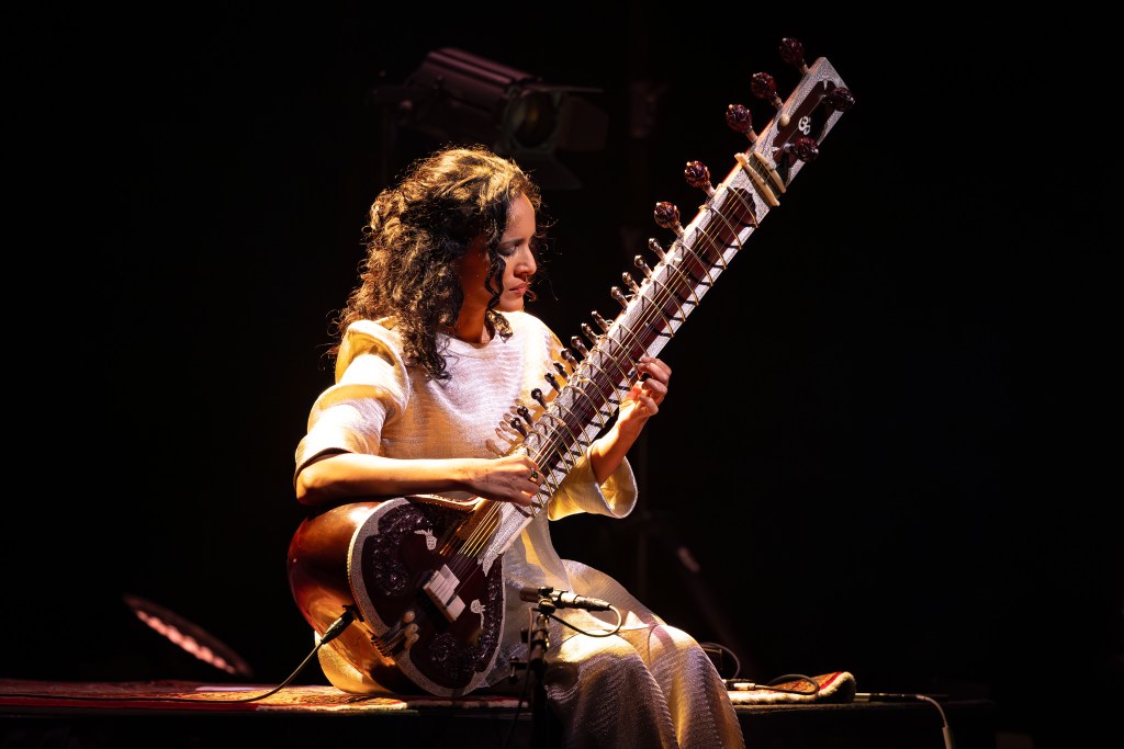 Anoushka Shankar. Woman sitting playing sitar against a black background.