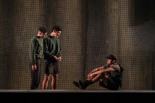 Saplings. Image is three young men on stage, one seated on the ground, two standing and looking down at him. They are all behind a large net-like curtain.
