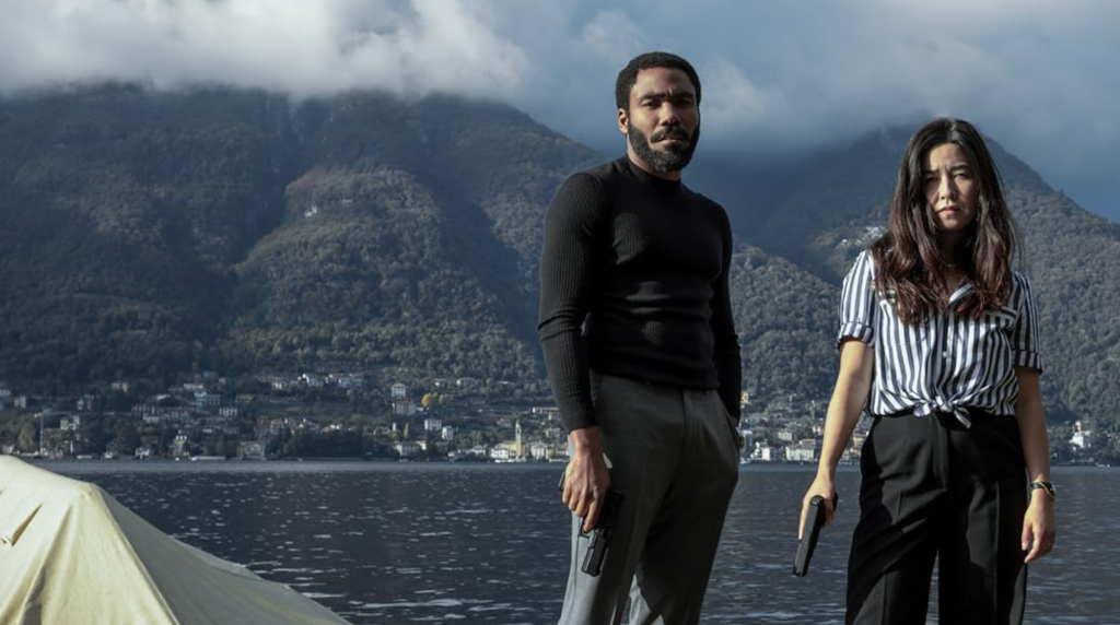 A man and a woman holding guns are on a hill with a lake and another mountain in the background.