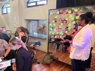 Cessalee Stovall delivers a Stage A Change workshop at Darlinghurst Theatre Company. Photo: Amylia Harris. Stovall stands on the right hand side, speaking to a group of people who are sitting around tables. She is a woman with black curly hair tied up in a bun, brown skin, wearing a pink cardigan.