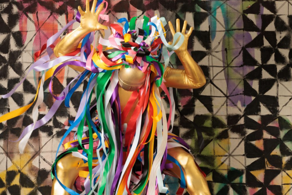 2024 visual arts. Image is gold coloured person with legs bent and hands in the air draped in multicoloured streamers.