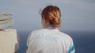 The Theatre of War. Image is a young woman in a white jacket with a blue stripe around the arm, hair in a bun, shot from the back and looking out to sea.