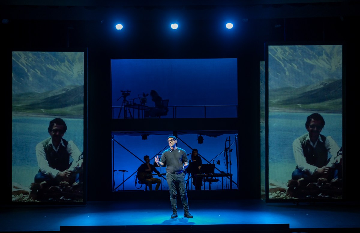 ‘Things Hidden Since the Foundation of the World’. Photo: Wendell Teodoro. A stage set with a single performer standing between two large-scale projection screens. The stage is lit blue.