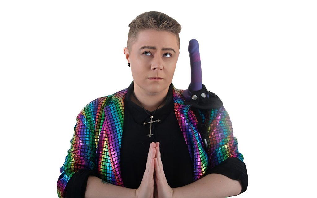 'A Night with Pastor Ace' by Ace R L. Image: Supplied. A non-binary person standing with their hands held in prayer, wearing a rainbow blazer and a dildo with googly eyes on their shoulder.