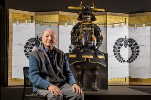 Vale. Max Carter. Aged man sitting with hands on knees and art behind him.