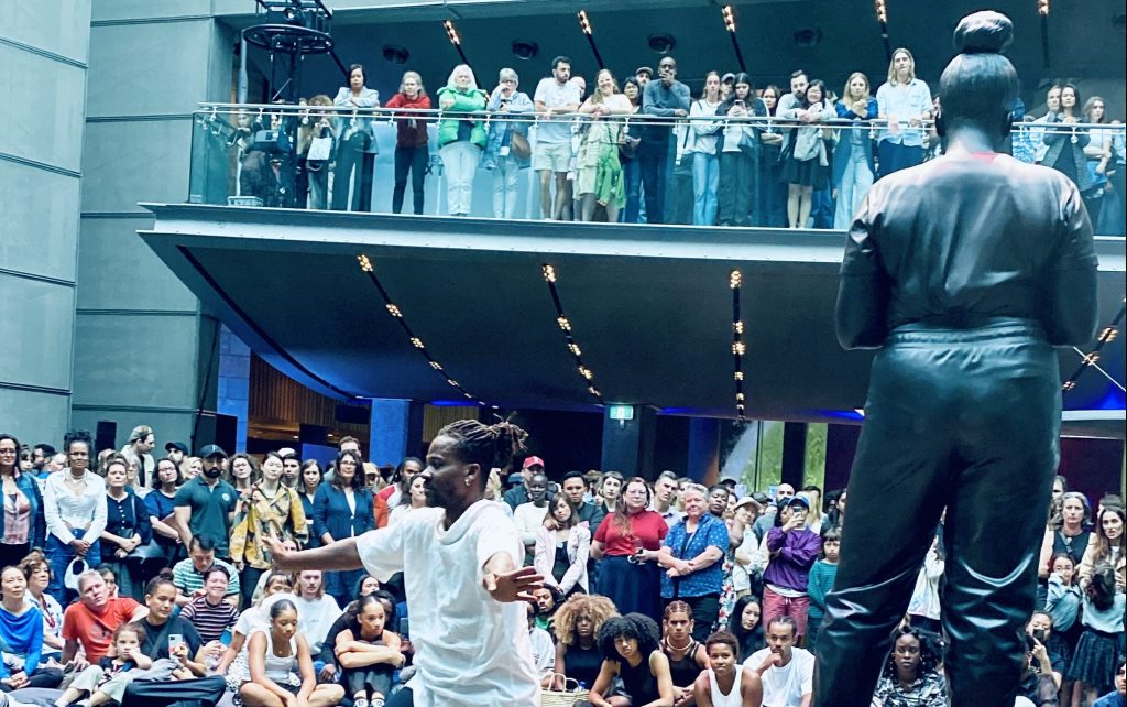 Afrocentric. Image is black dancer, dressed in white performing in gallery space at front of the NGV, beside a large black sculpture of a young black woman. There is audience seated around the space.