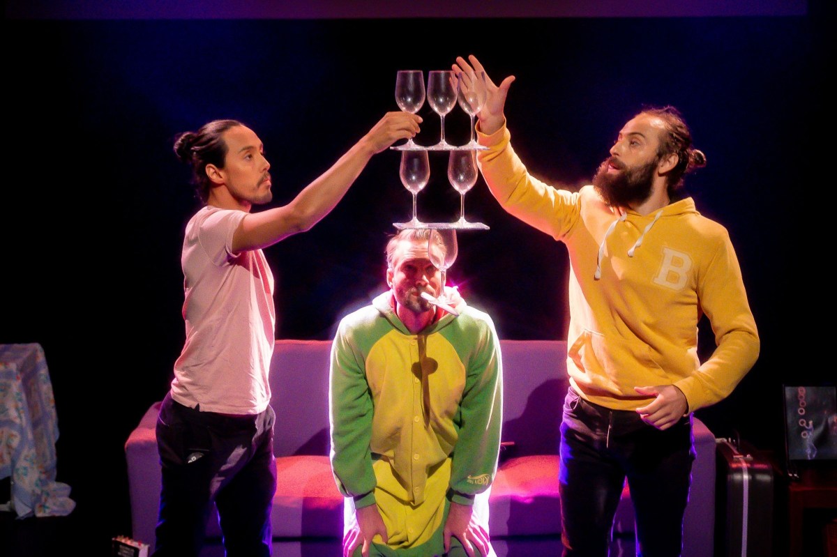 Appeasing Nergal. Three performers attempting to balance wine glasses in front of a sofa against a black backdrop.