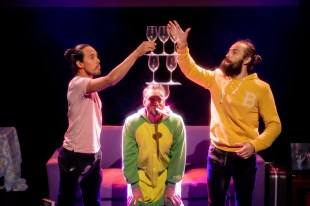 Appeasing Nergal. Three performers attempting to balance wine glasses in front of a sofa against a black backdrop.