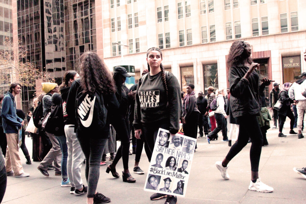 Australia has a black history. Image is a photograph of a group of black clad protesters in the streets of Sydney. A young woman with braided hair, a Black Lives Matter t shirt and a placard is in the centre.